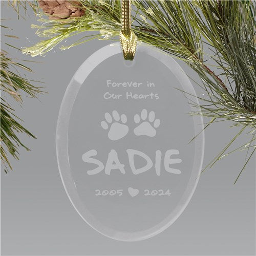 Clear glass ornament with engraved text and paw prints, hanging on a Christmas tree branch.