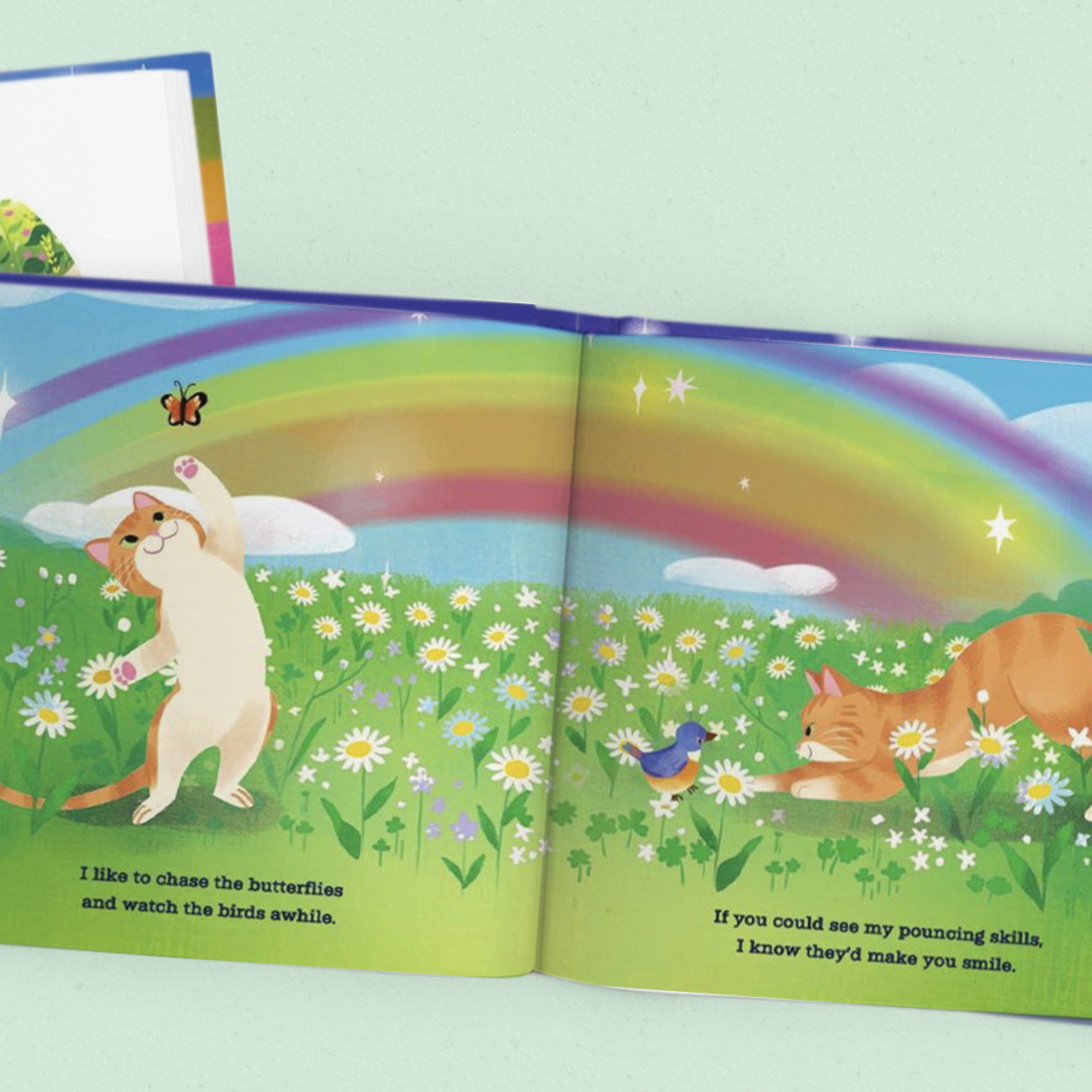 Open children's book with cartoon cats in a field and rainbow on a light green background