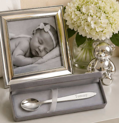 Silver spoon and letter opener set with engraved name, framed photo of a baby, and white flowers.